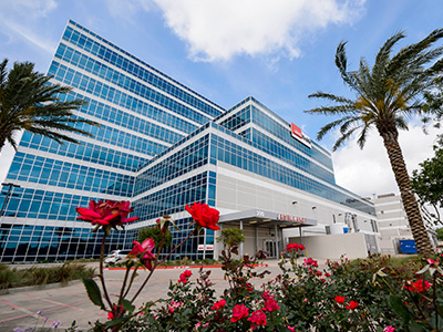 Exterior of UTMB Clear Lake campus with glass windows, palm trees and rose gardens.