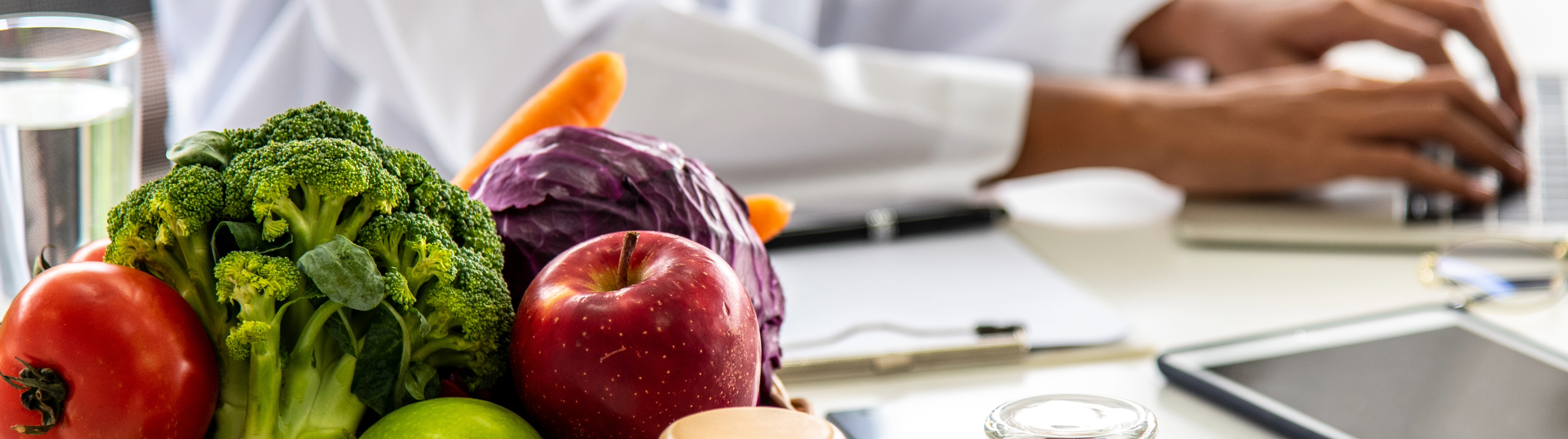 close up image of a tomato, broccoli, apple, cabbage and carrot with a pair of hands typing on a laptop in the background