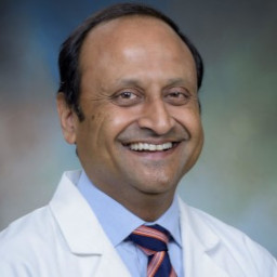 Formal studio portrait of a UTMB Innovation and Entrepreneurship faculty member wearing a white lab coat, dress shirt, and tie against a soft blue‑green background.