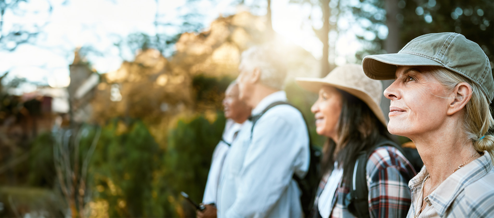 Older adults hike outdoors, smiling and enjoying nature. A woman in front, in a cap and plaid shirt, looks ahead with a content expression. 