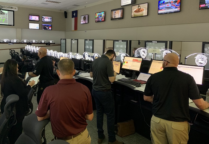 Personnel stand at computer workstations in a command center with multiple monitors and wall-mounted screens displaying maps and news feeds.
