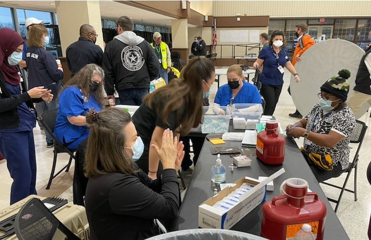 Staff at tables assist individuals with registration and medical services, with supplies, paperwork, and safety equipment arranged indoors.