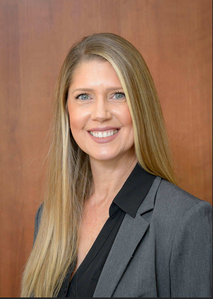 Professional headshot of Tilly Clark, Director of Presidential Affairs and Event Planning at the University of Texas Medical Branch, wearing a gray blazer and dark blouse, photographed against a warm wood‑tone background.