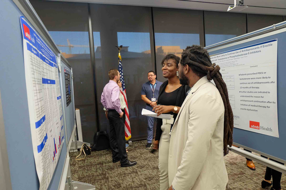 Two students stand beside research posters during a doctoral poster session while other attendees talk in the background.
